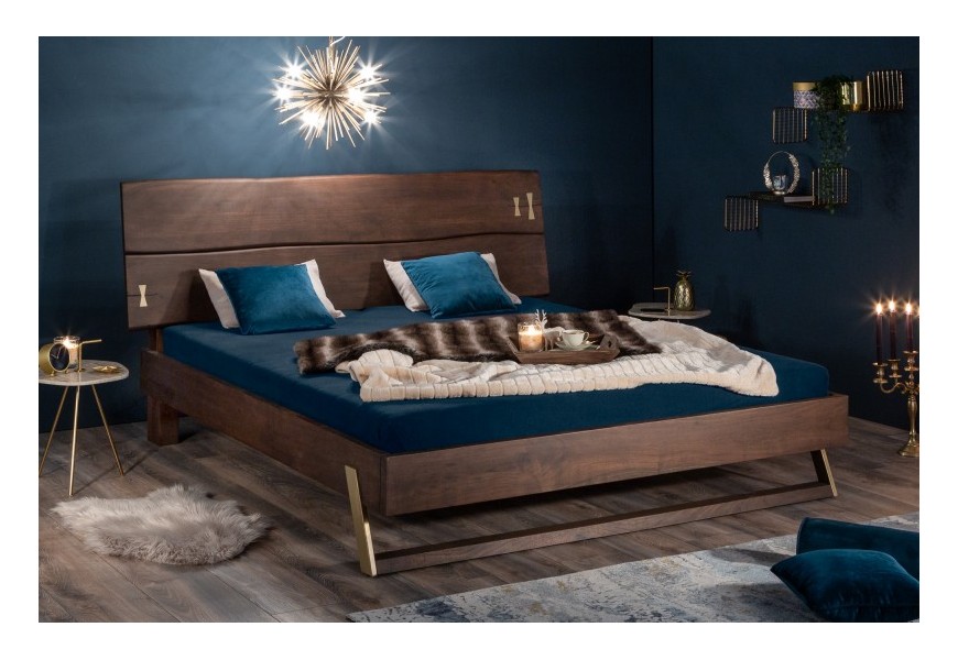 Štýlová masívna posteľ Mammut z dreva Akácie doplnená zlatými dekoráciami na zvlnenom čele a atypickej lyžinovej nohe
