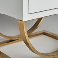 Moderný biely nočný stolík Bynum v sklenenom vyhotovení s dvoma zásuvkami a zaoblenou kovovou podstavou v zlatej farbe 60cm