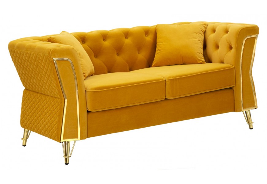 Dizajnová art deco žltá čalúnená dvojsedačka Velaria s poťahom zo zamatu s dekoratívnym prešívaním na opierke v chesterfield štýle a diamantovým vzorom na vonkajšej strane s dvomi vankúšmi a dekoratívnym zlatým prvkom na opierkach a šikmými nohami z kovu v zlatej farbe